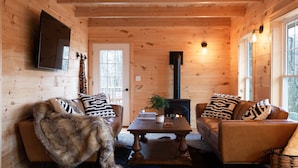 Ein gemütliches Blockhauskaminzimmer mit Holzwänden, einem Kamin, zwei Ledersofas mit gemusterten Kissen, einem Couchtisch und einem Wand-Fernseher.