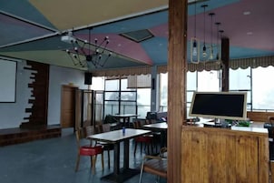 Un área de comedor con mesas y sillas, una barra de madera con un monitor de computadora, y una escalera que lleva a otro nivel.