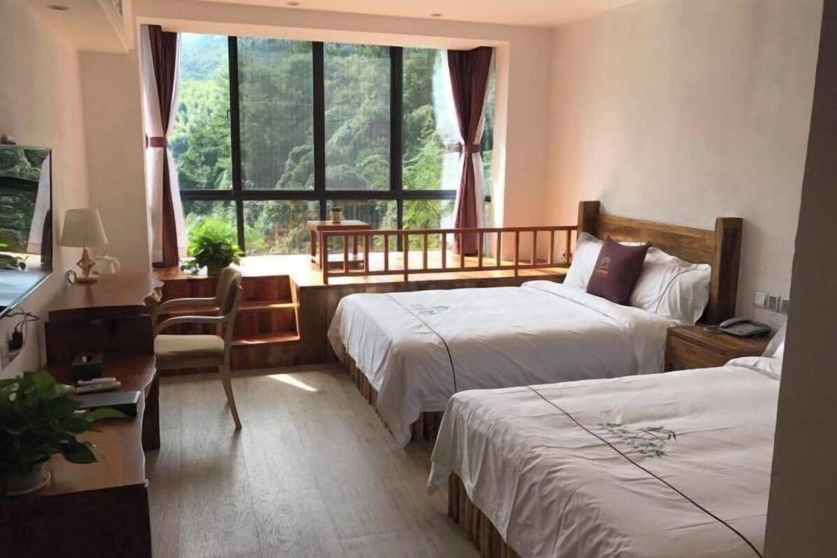 Habitació d'hotel amb dos llits, un escriptori amb una làmpada, una cadira i vistes a la forest a través de grans finestres.