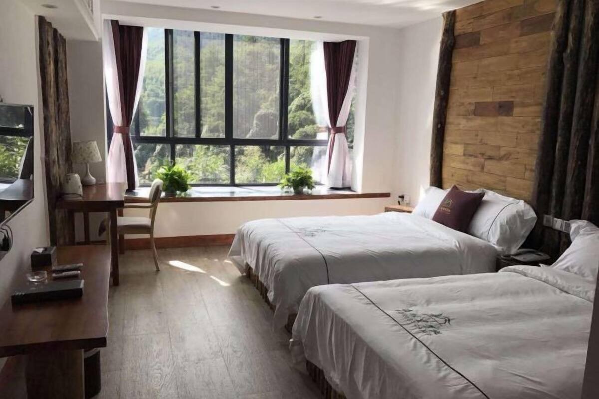 Hotellihuone, jossa on kaksi sänkyä, puinen työpöytä, ikkuna verhoilla ja näkymä vehreään maisemaan.