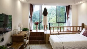 A bedroom with a bed, a desk, a chair, a TV, and a window with a view of greenery. - Senlinli Campsite (Zixing)