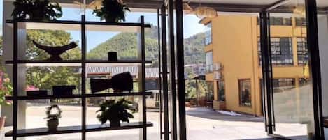 A room with a glass door leading to an outdoor area, shelves with potted plants, and a view of a building and hills.
