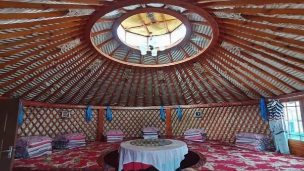 A circular yurt with a central dome, a bed with a red and white patterned bedspread, and a small table with a white cloth.