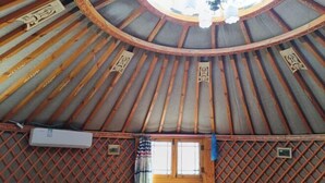 A yurt with a circular wooden ceiling, a bed with a patterned bedspread, a window with curtains, and a small air conditioning unit.