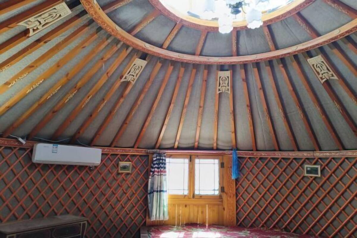 A yurt with a circular wooden ceiling, a bed with a patterned bedspread, a window with curtains, and a small air conditioning unit.