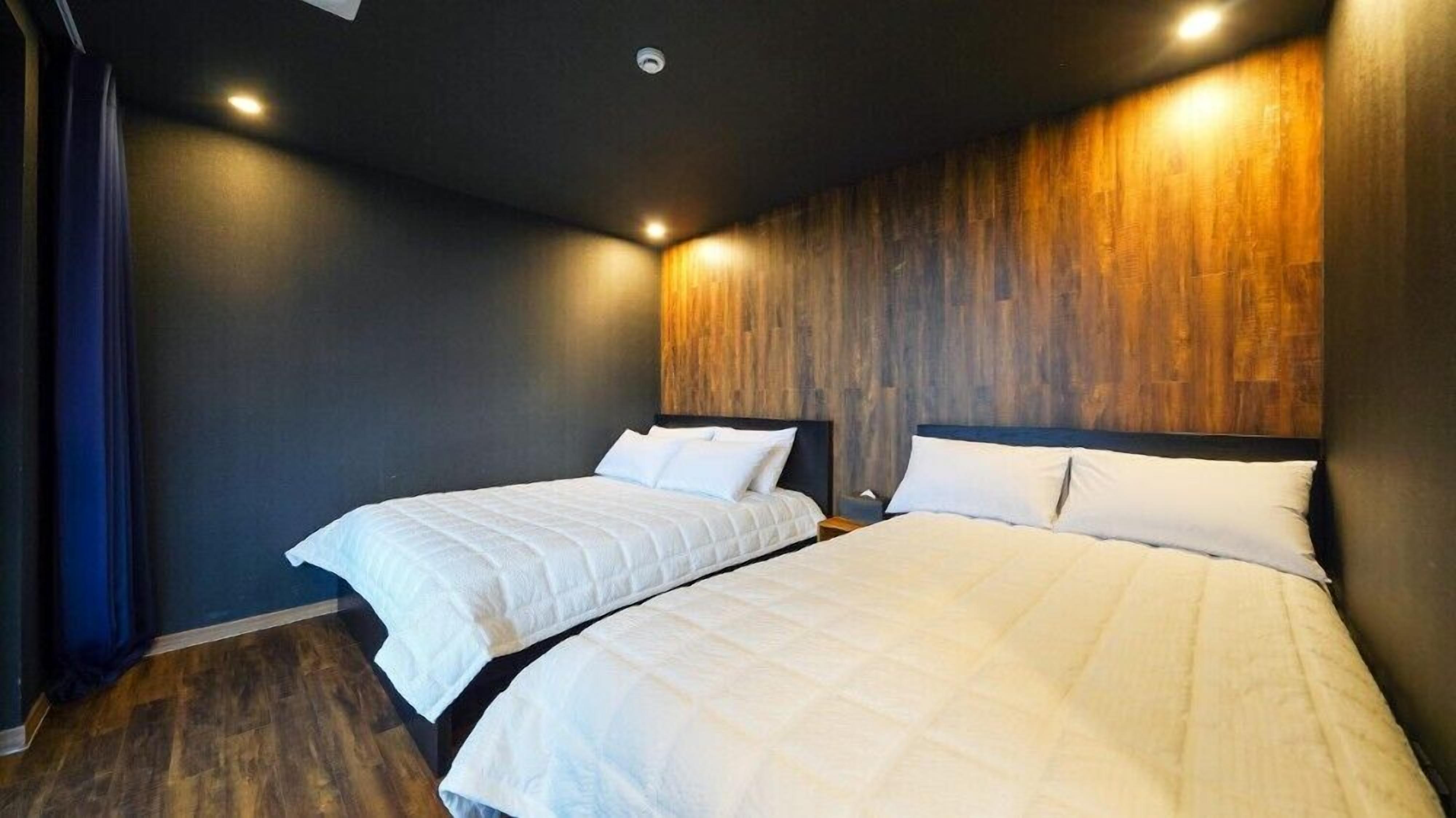 Habitación de hotel con dos camas, paredes oscuras, paneles de madera en una pared y una lámpara en el techo.