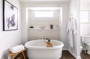 Un baño moderno con una bañera independiente, un taburete de madera pequeño, una toalla, un lavabo y una ventana sobre el mueble de tocador.