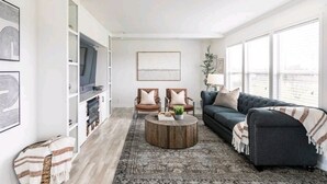 Un salón espacioso con un ventanal grande, un sofá de color gris oscuro, dos sillones, una mesa de centro de madera y un televisor sobre un mueble blanco.