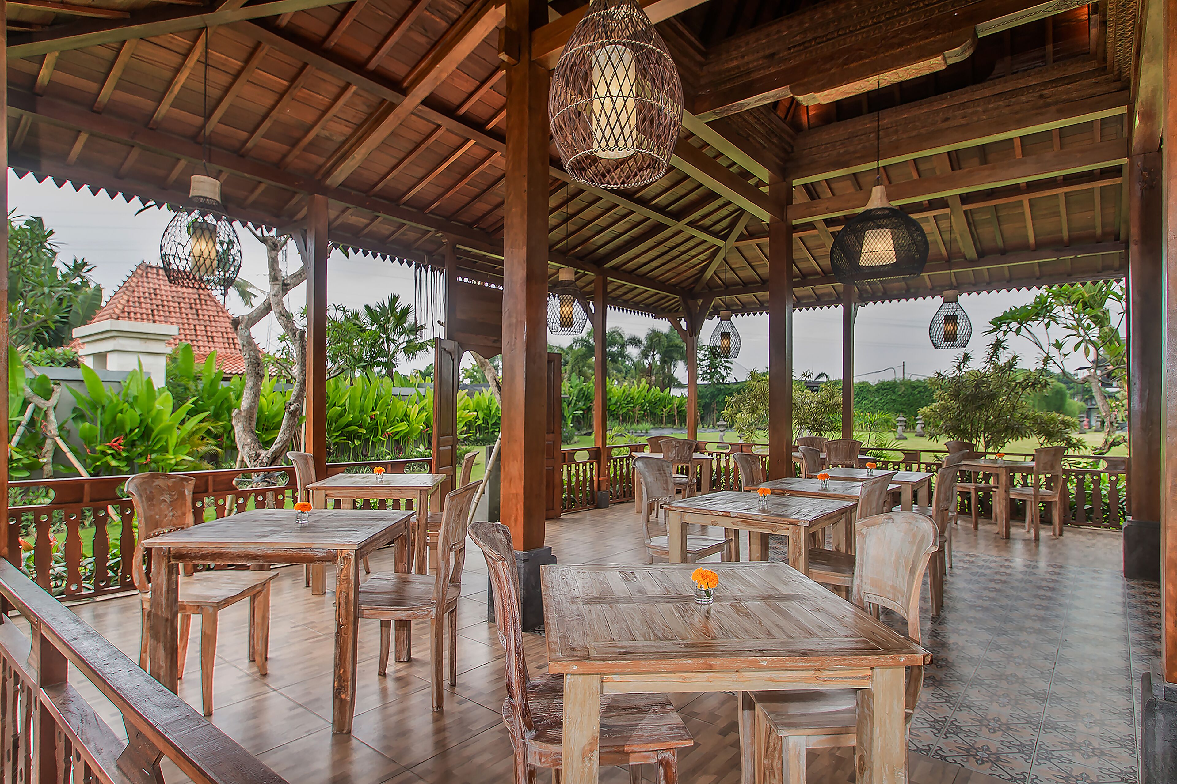 An outdoor wooden pavilion with tables and chairs, surrounded by lush greenery and a view of a traditional building.