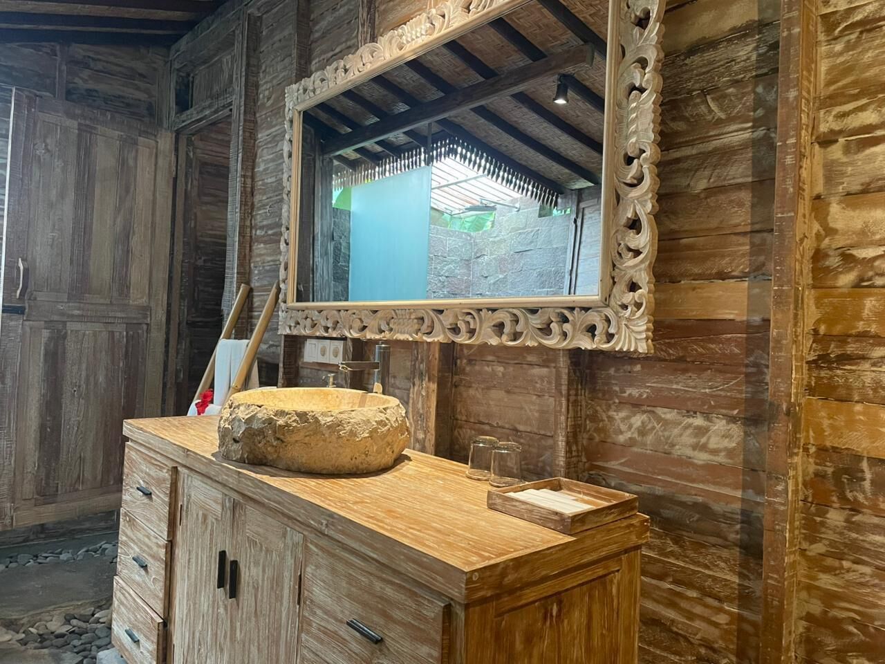 A rustic bathroom with a wooden vanity, a large mirror with ornate carvings, and a stone basin.