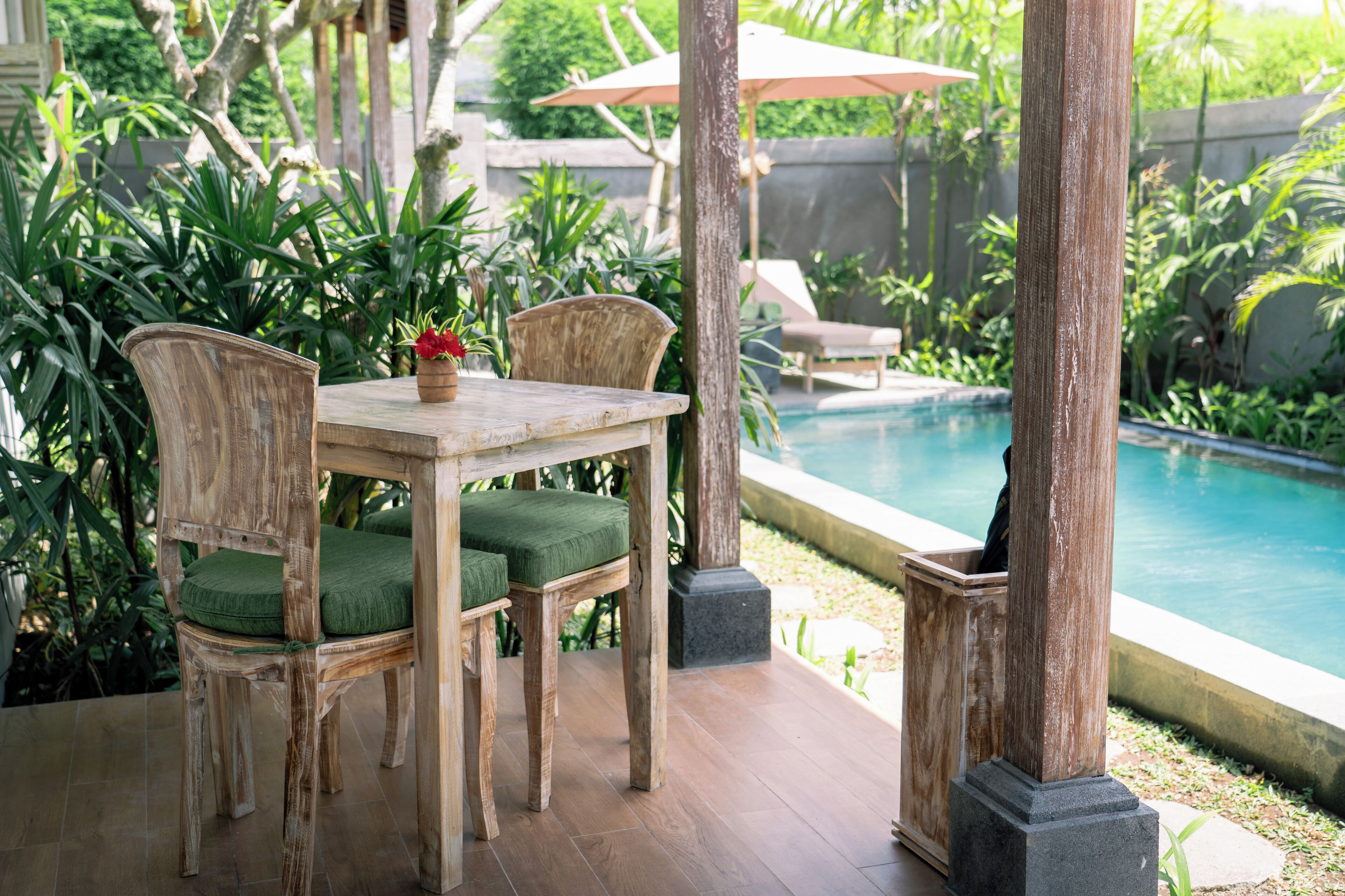 A wooden outdoor dining area with a table and chairs, a small potted plant, and a view of a swimming pool.