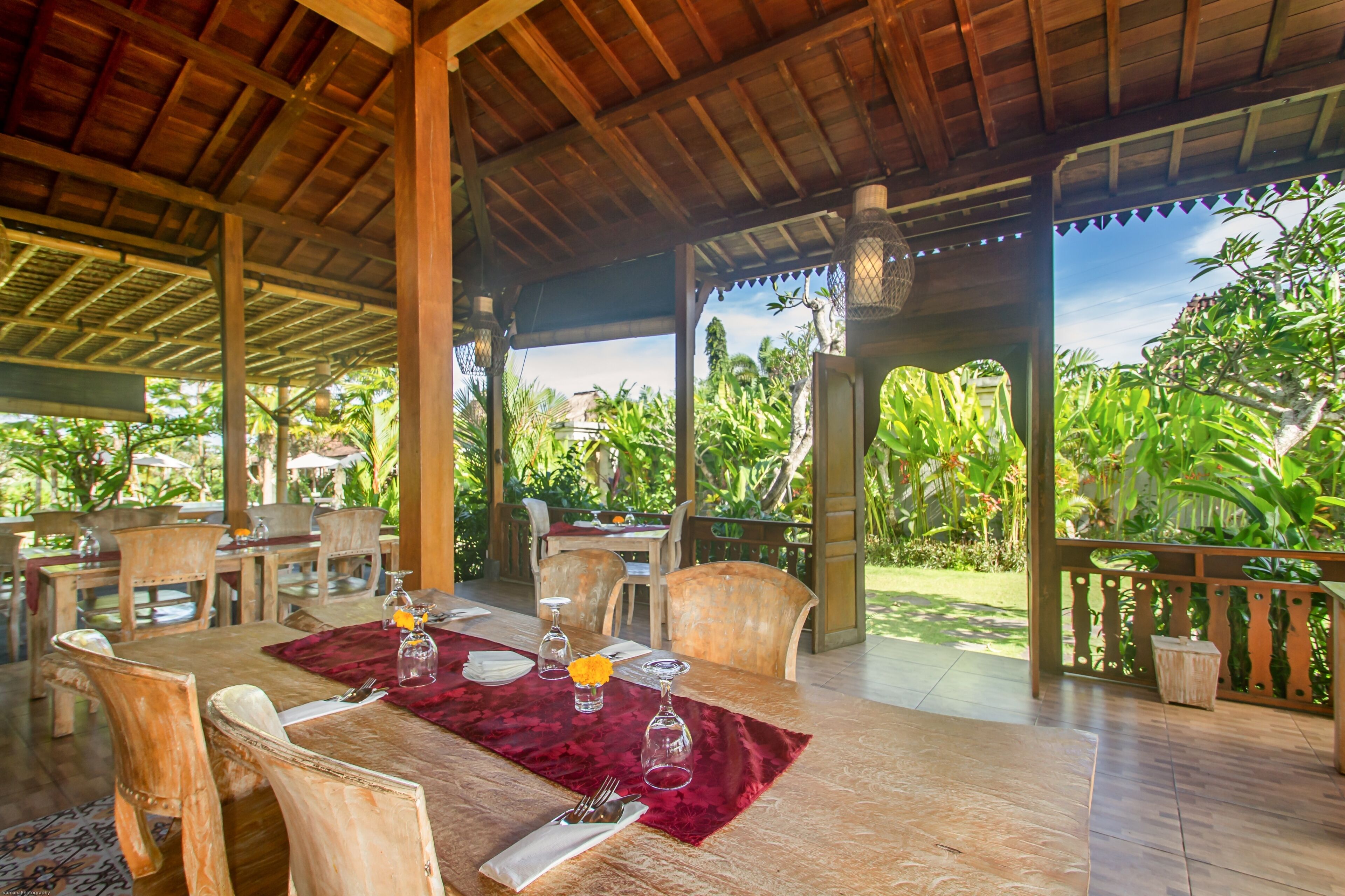 An outdoor dining area with wooden tables and chairs, a red table runner, and a view of lush greenery.