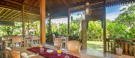 An outdoor dining area with wooden tables and chairs, a red table runner, and a view of lush greenery.
