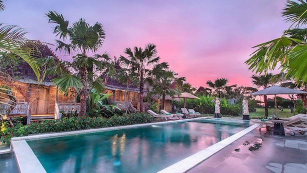 A swimming pool with lounge chairs and palm trees, surrounded by a traditional building with a thatched roof.