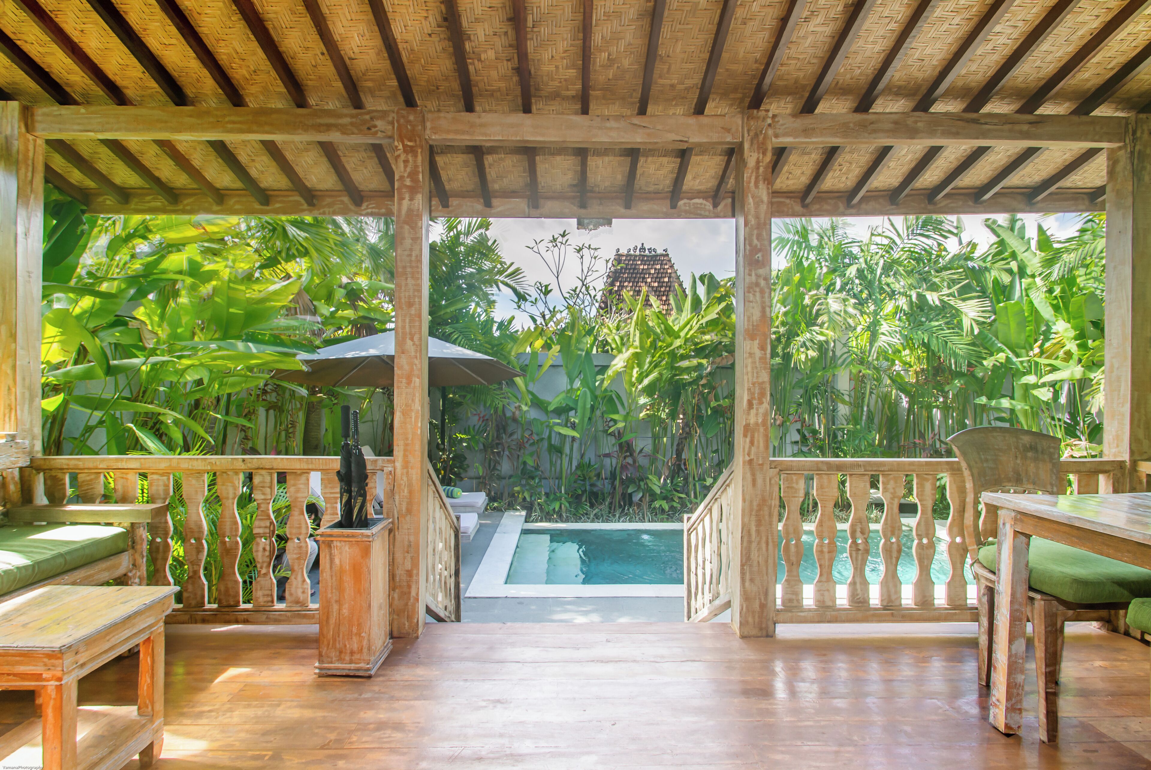 A wooden deck with a pool, surrounded by lush tropical plants and a view of a traditional building.