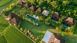 An aerial view of a resort with multiple thatched-roof cottages, a central swimming pool, and lush greenery.