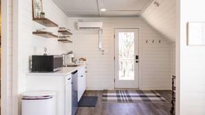 Cocina compacta con armarios blancos, microondas y cafetera. Hay una puerta que da a un balcón con vistas a árboles.