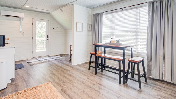 Una habitación amplia con suelo de madera, una zona de comedor con mesa y taburetes, una puerta con ventanal e iluminación empotrada en el techo.