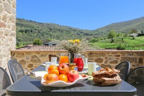 A table with fruits, a vase with flowers, and drinks on a terrace with a stone wall and a view of green hills.