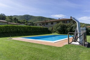 A rectangular swimming pool with a wooden deck, surrounded by well-maintained grass, a stone house with a tiled roof, and a hilly landscape in the background.
