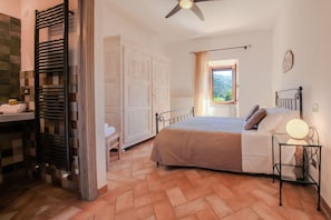 A bedroom with a bed, a bedside table with a lamp, a window with a view, and a door leading to another room. - Villa Fornace by MMega (Lisciano Niccone)
