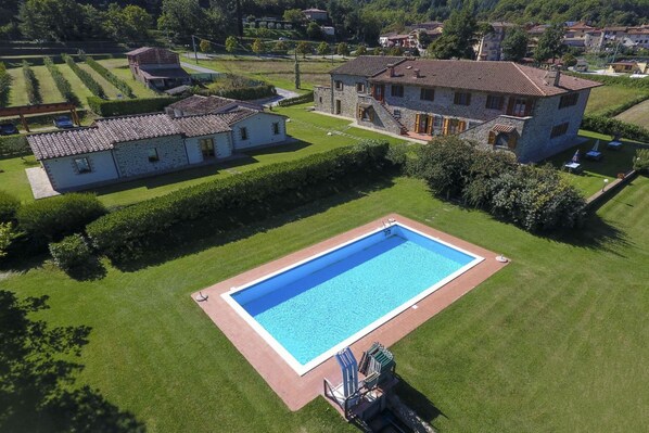 A large rectangular swimming pool with a surrounding deck, adjacent to a stone building with a tiled roof and a smaller white building with a tiled roof.