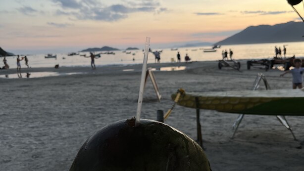 A hand holding a coconut with a straw, set against a beach scene with people and boats in the background. - House on a BUS by the river (Florianópolis)