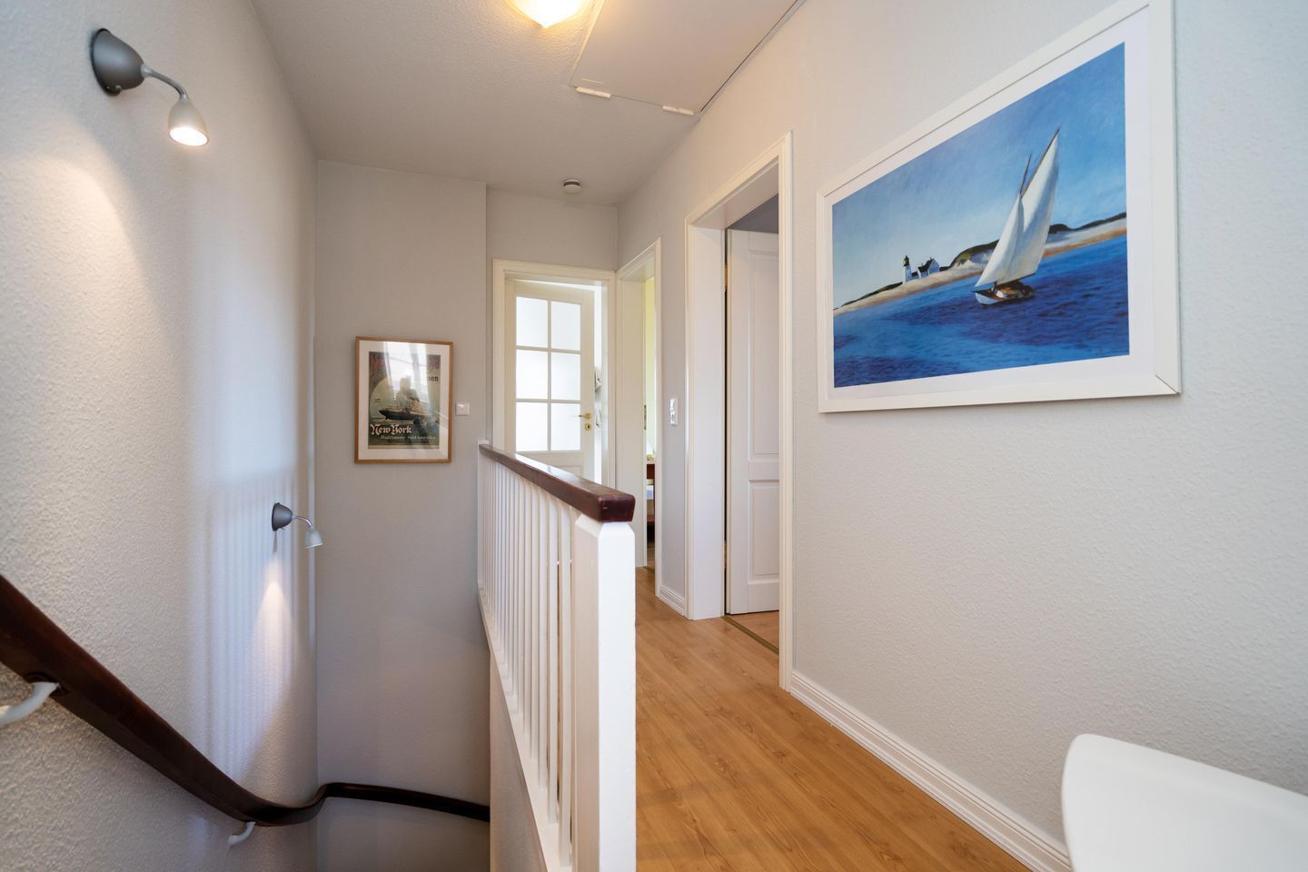 Un couloir doté d’un plancher en bois, d’une porte blanche, d’un tableau encadré représentant un voilier et d’un luminaire fixé au mur.
