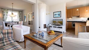 Una sala de estar moderna con una mesa de centro de vidrio, sofás blancos y una zona de cocina con armarios de madera y una mesa de comedor.
