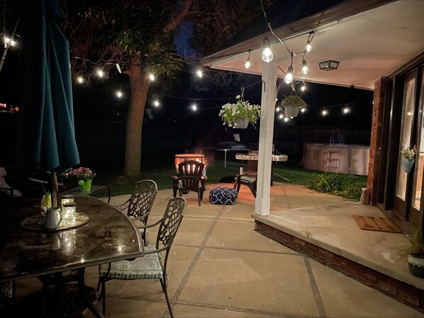 Un patio al aire libre con una mesa redonda, sillas, plantas colgantes y luces de cuerda.