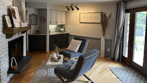 Un salon moderne avec une cheminée, un coin repas et une cuisine. Il y a une grande fenêtre avec des rideaux, un luminaire fixé au mur et une pancarte sur laquelle on peut lire : « Voici notre famille, unie et comblée. »