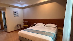 A hotel room with a large bed, a wooden headboard, a bedside table with a lamp, a chair, and a wall-mounted painting. - TD CAPITAL HOTEL (Kota Kinabalu)