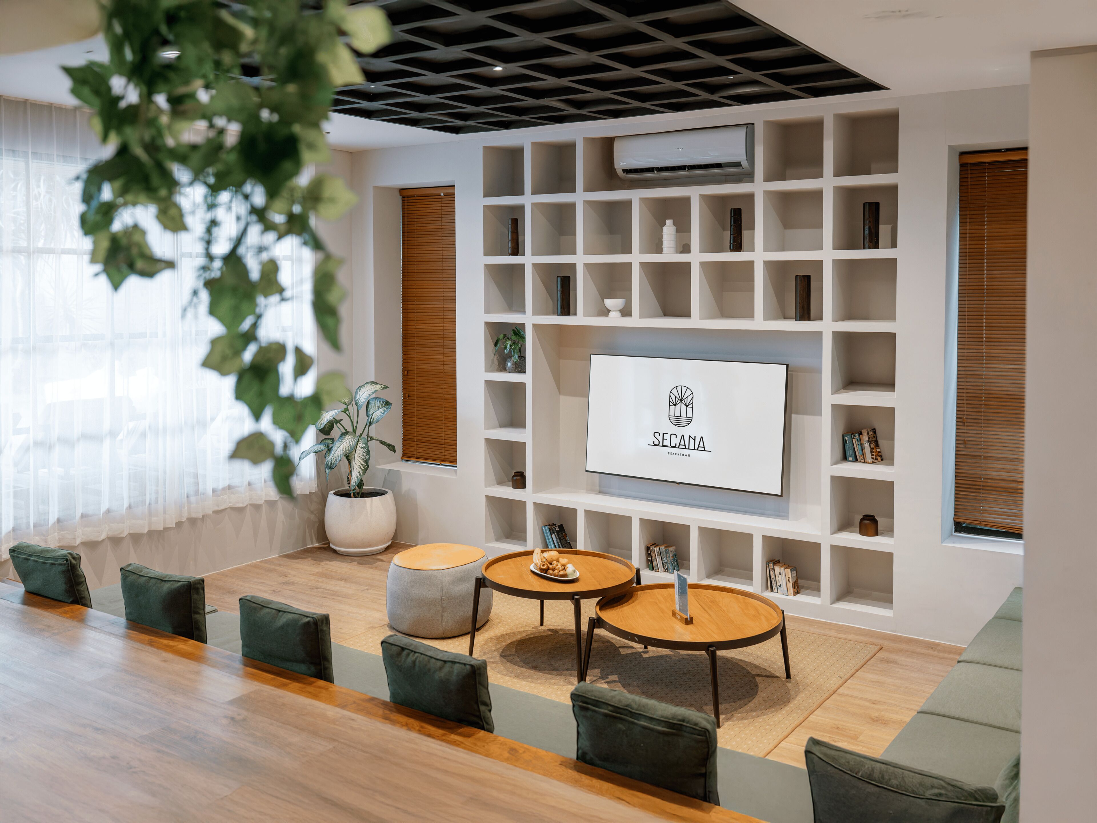 A modern interior with a wooden floor, green upholstered chairs, a round wooden table, a white wall-mounted TV, and a potted plant.