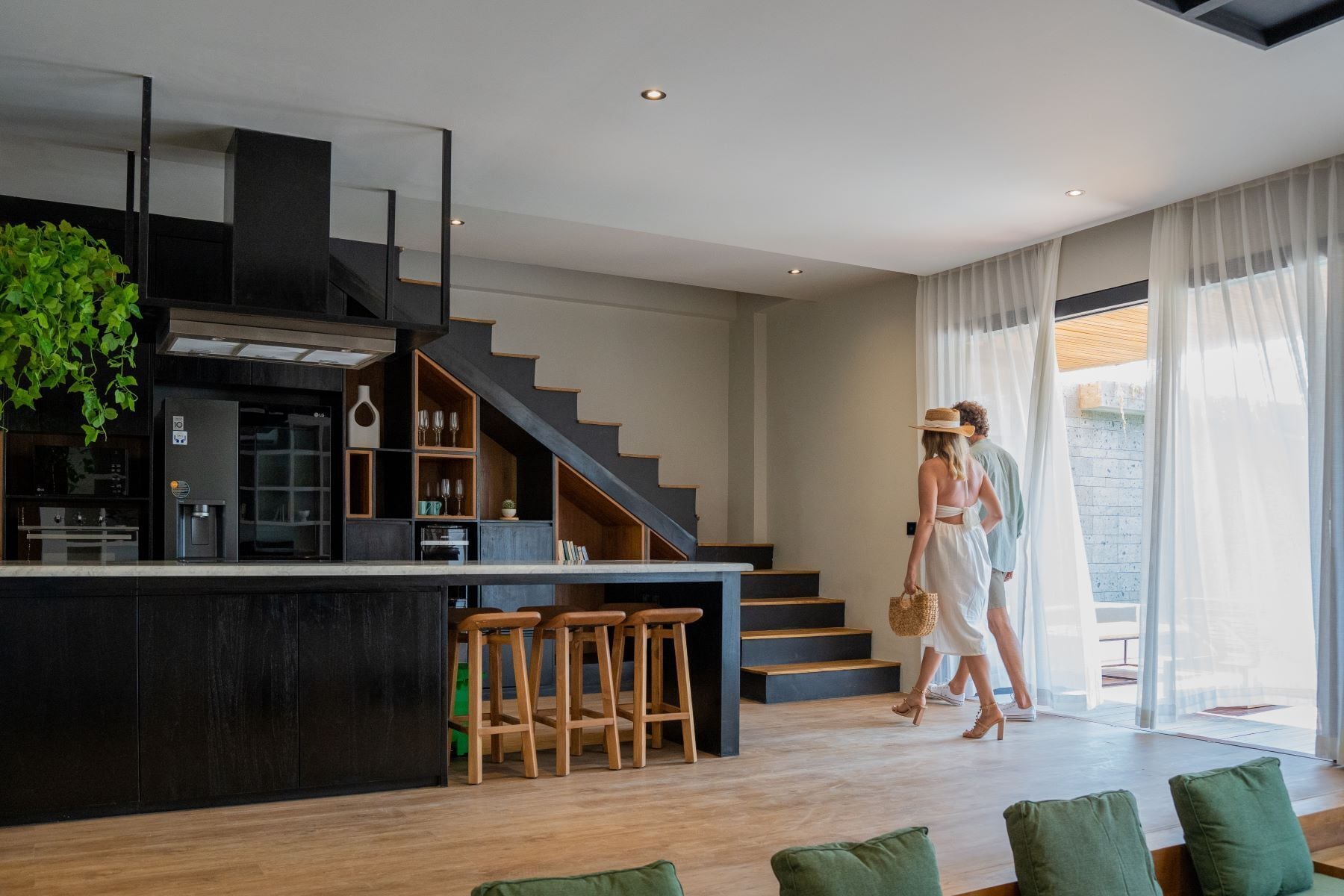 A modern kitchen with a black countertop, wooden stools, and a staircase leading to a balcony.