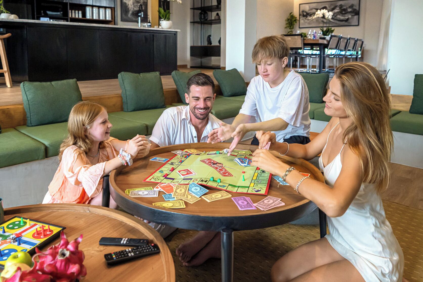 Un groupe de personnes joue à un jeu de société dans un salon moderne, avec des canapés verts et une table basse en bois.