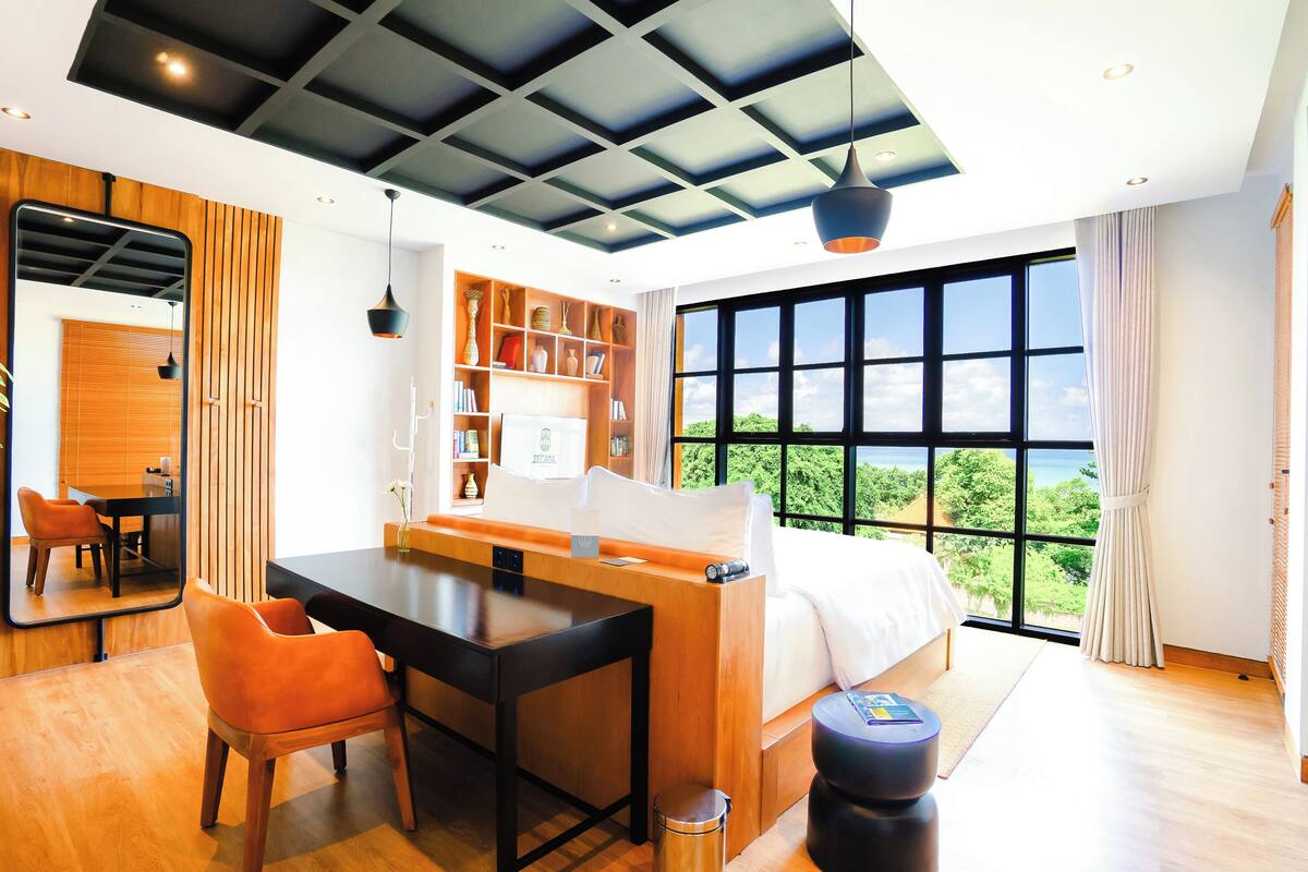 Habitación de hotel moderna con una cama grande, un escritorio y una zona de estar. Se aprecia vista al exterior a través de amplios ventanales.