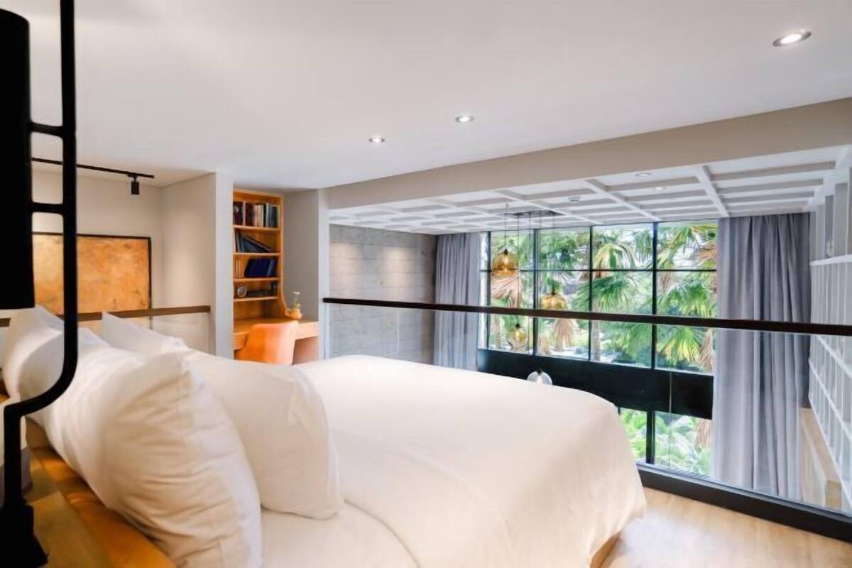Habitación de hotel moderna con una cama grande, una estantería y vistas a palmeras a través de amplias ventanas.