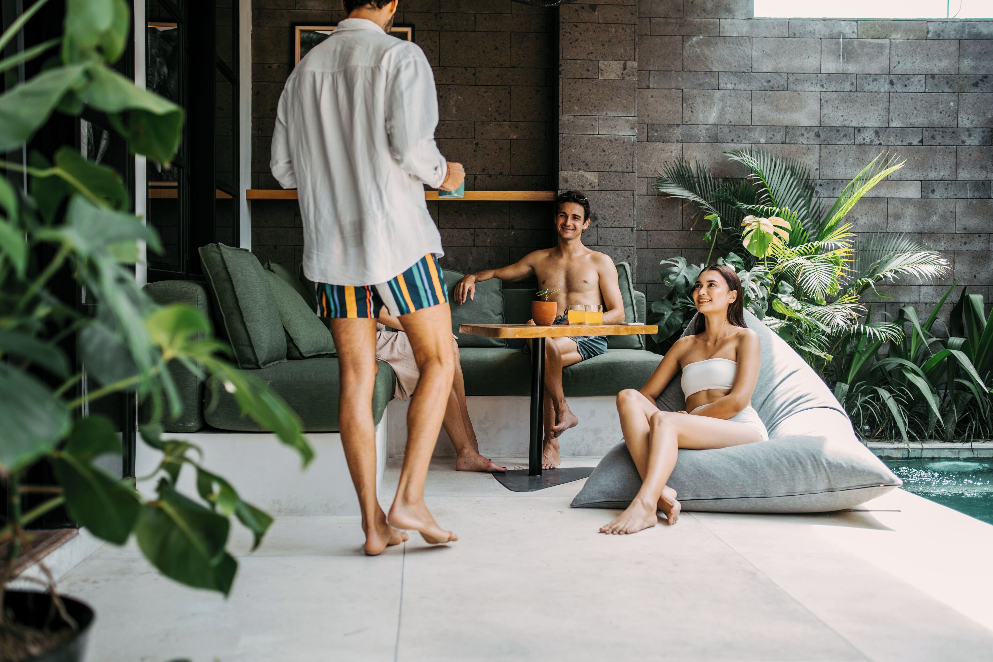 A modern indoor pool area with a green sofa, a dining table with a drink, and a person standing.