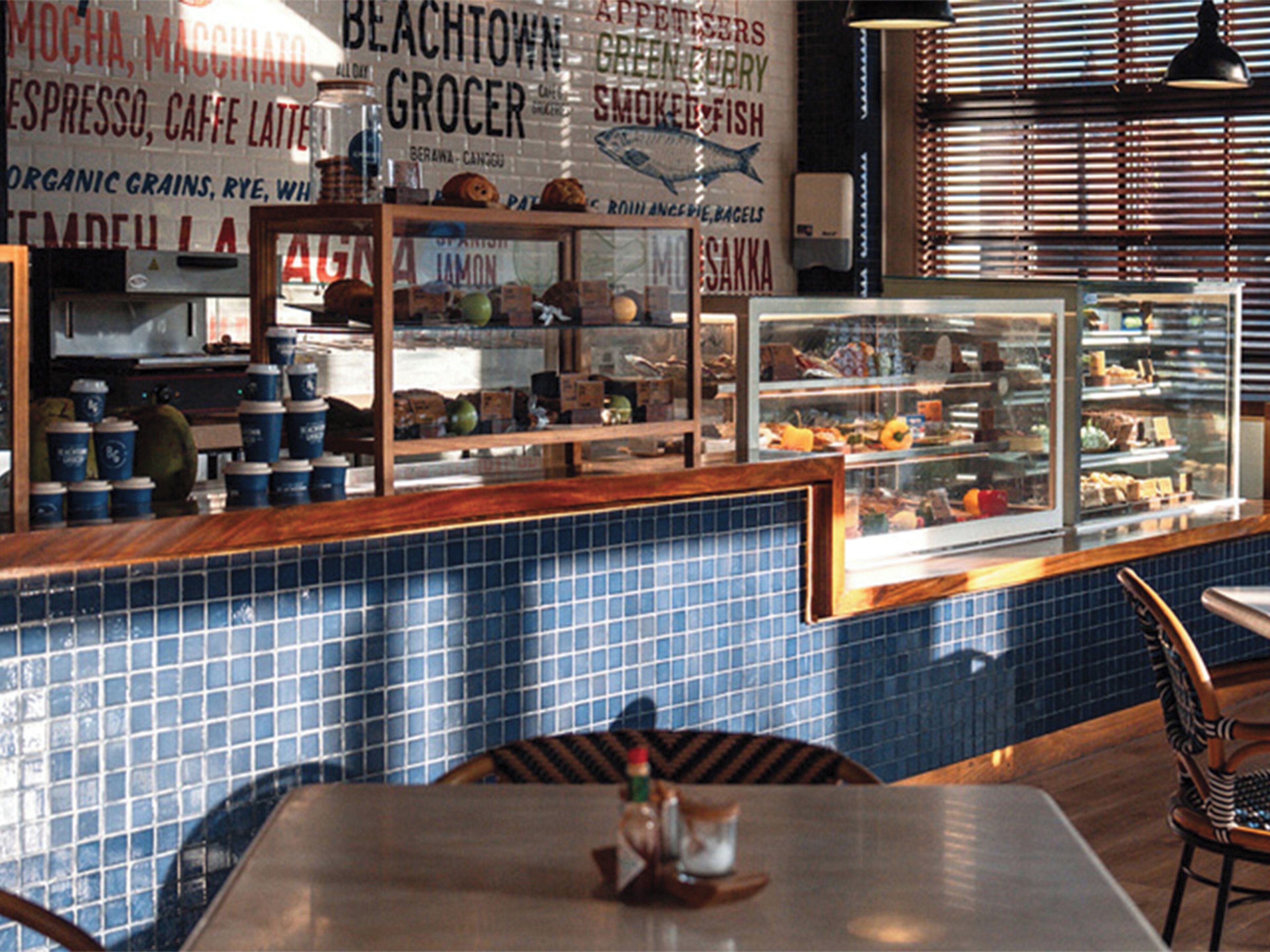 A cafe interior with a counter displaying coffee cups, a glass display case with pastries, and a blue tiled backsplash.