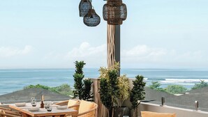 Un espacio para comer al aire libre con sillas y mesas de mimbre, un alto pilar con una planta y vista al océano.