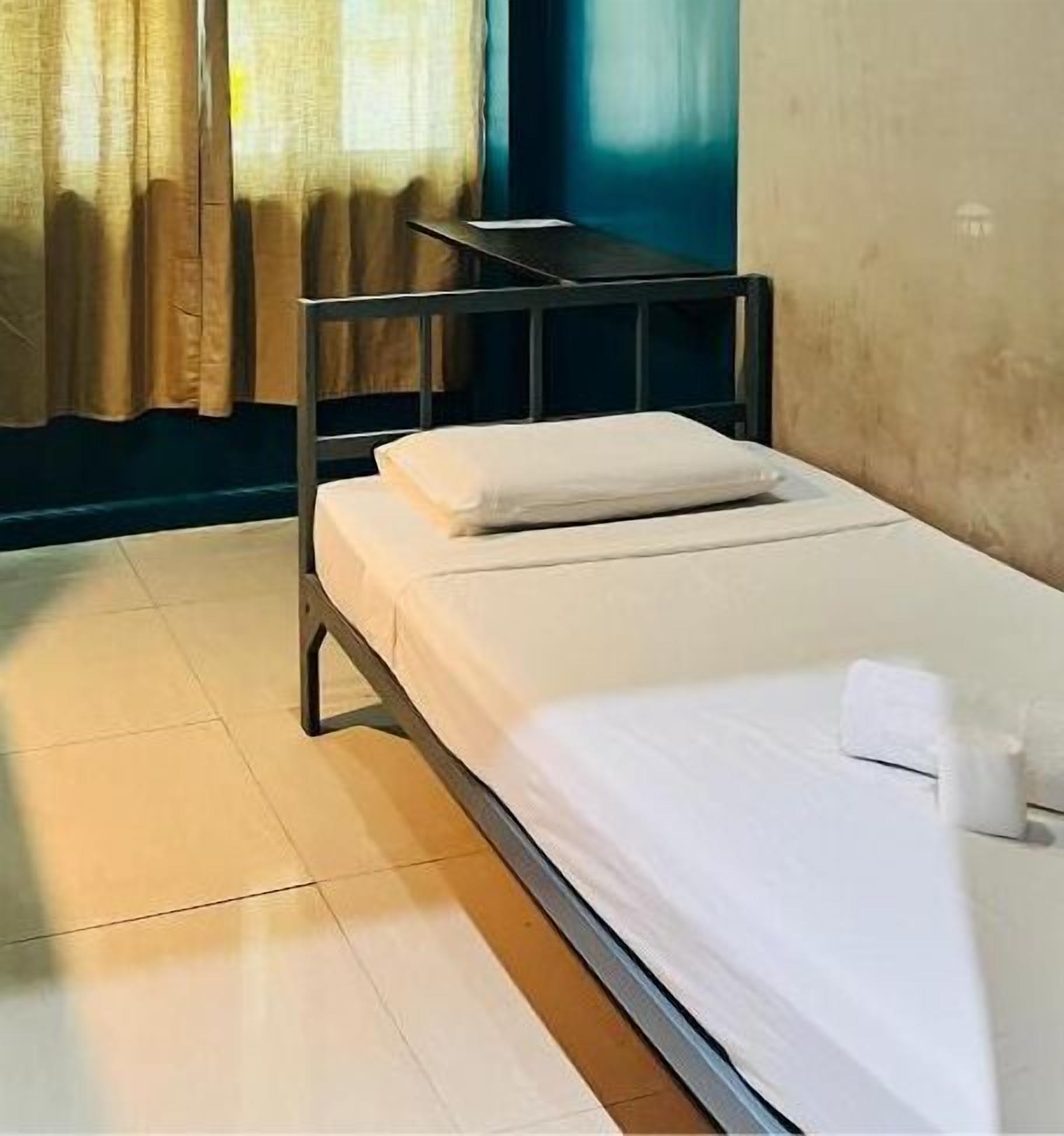 A single bed with a white mattress and pillows, a wooden headboard, a small table, and a window with curtains.