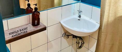 Um banheiro com uma pia simples, um espelho e uma prateleira com a etiqueta 'SINGLE BASIC AMENITIES'.