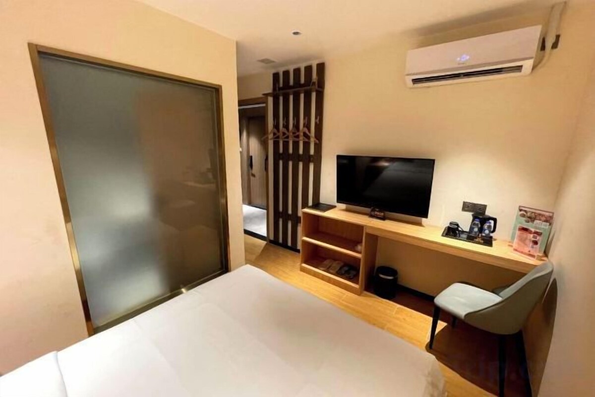 A hotel room with a bed, a TV, a desk with a chair, and an air conditioning unit.