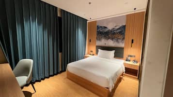 A hotel room with a large bed, a desk, a chair, a nightstand with a phone, and a wall with a mountain painting.