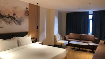 A hotel room with a bed, a sofa, a small table, a desk with a lamp, a painting on the wall, and a window with curtains.