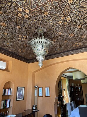 A room with a patterned ceiling, a chandelier, bookshelves, framed pictures, and a dining area.