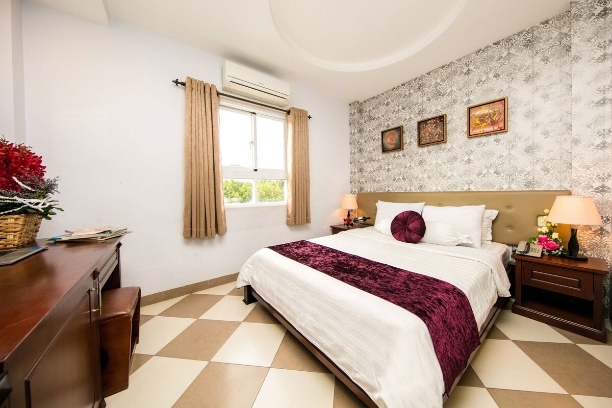 Habitación de hotel con cama, escritorio, silla, ventana con cortinas, pared con cuadros enmarcados y papel tapiz estampado.