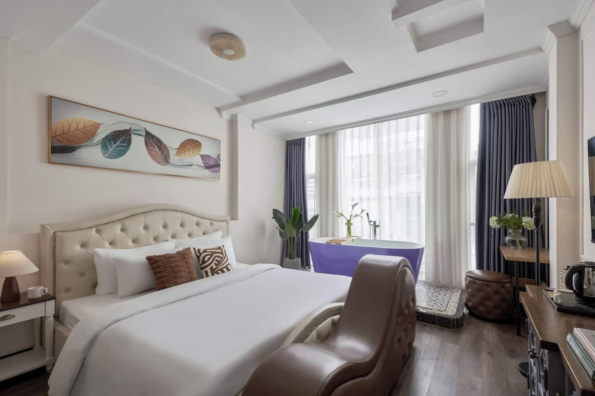 Ein Hotelzimmer mit einem großen Bett, einem Sessel, einem Schreibtisch, einem Tisch und einem Vorhangfenster.