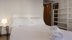 A hotel room with a bed, a nightstand with a lamp, a wooden door, and a white shelving unit. - Chianti's Inn Trebbiano (Firenze)