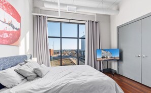Quarto com uma cama grande, uma televisão sobre um suporte e vista para a paisagem urbana através de uma janela ampla.